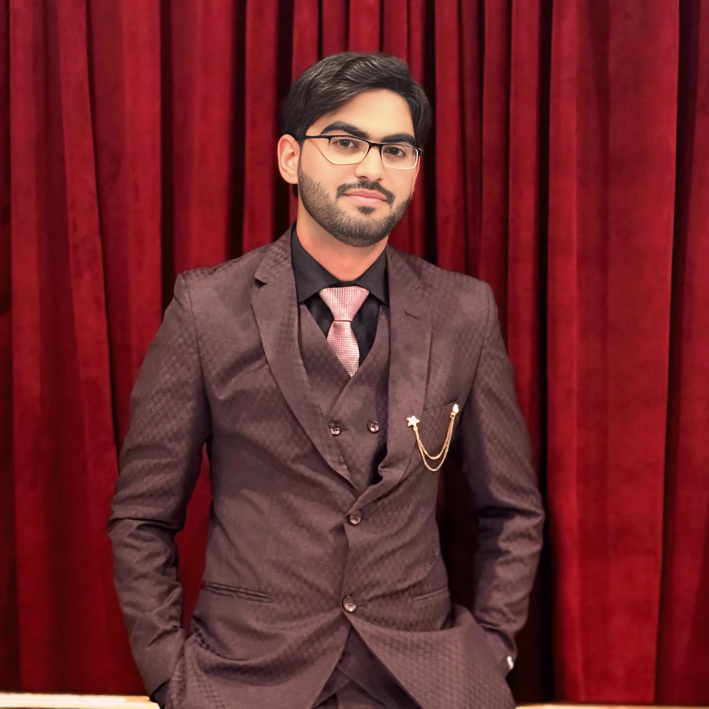 Dr. Faheem Ghaffar wearing a formal maroon suit with a pink tie standing in front of a red curtain.