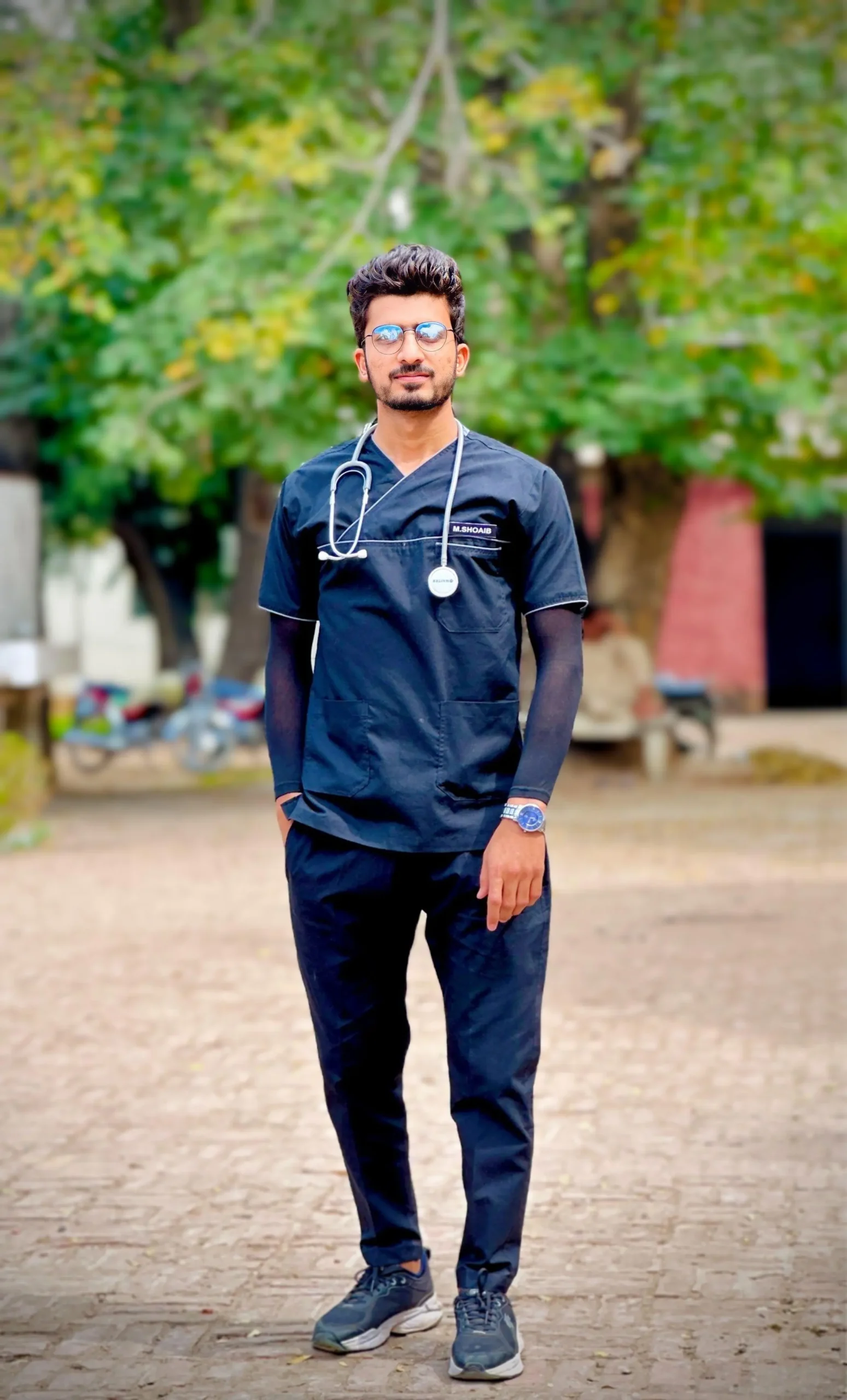 Dr. Shoaib Maqbool is standing outdoors wearing black scrubs and a stethoscope.